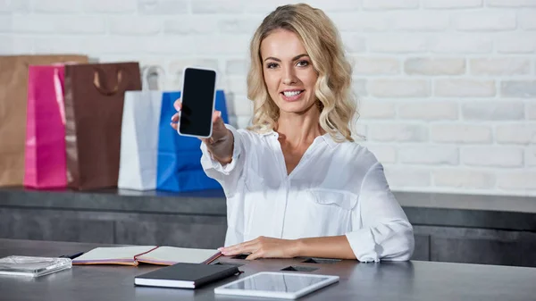 Beautiful Young Seller Holding Smartphone Blank Screen Smiling Camera Store — Free Stock Photo