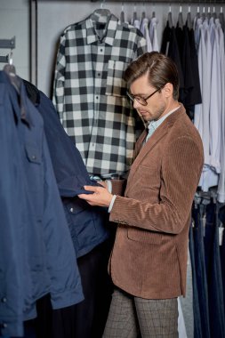 handsome man in eyeglasses looking at jacket while shopping in boutique     clipart