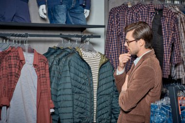 handsome young man with hand on chin looking at stylish clothes in boutique clipart