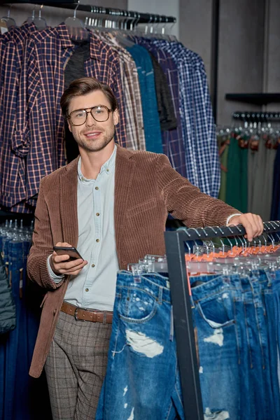 英俊的年轻男子在眼镜持有智能手机和微笑在相机在精品店 — 免费的图库照片