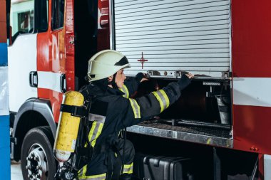 female firefighter with fire extinguisher on back closing truck at fire station clipart