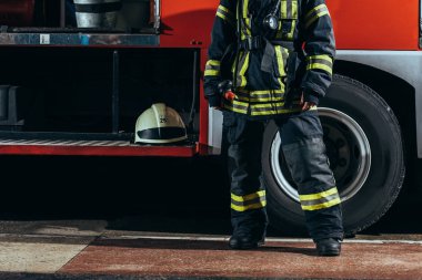 partial view of female firefighter in protective uniform standing near truck with helmet at fire station clipart