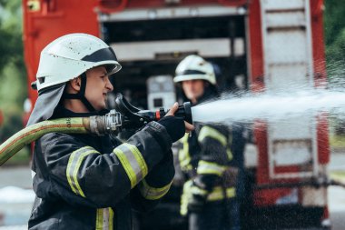 selective focus of firefighter with water hose extinguishing fire on street clipart