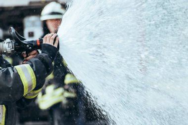 partial view of firefighter with water hose extinguishing fire on street clipart