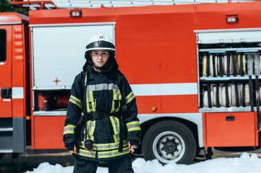 female firefighter in protective uniform standing on street with red fire truck behind clipart