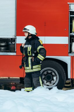 female firefighter in protective uniform standing in foam on street with red fire truck behind clipart