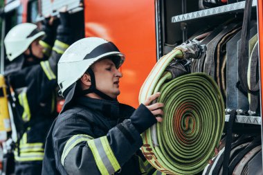 male firefighter in helmet putting water hose into truck on street clipart