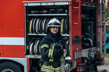 portrait of fireman in protective uniform and helmet standing at truck with water hoses inside on street clipart