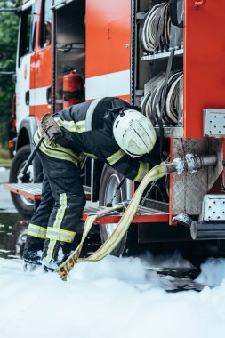 partial view of firefighter in protective uniform checking water hose on street clipart