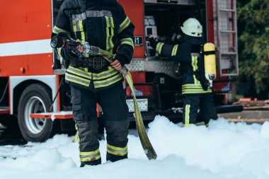 cropped shot of firefighter in water hose in hands standing in foam on street clipart