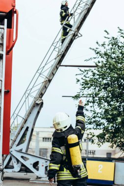 back view of female firefighter with fire extinguisher on back gesturing to colleague on ladder on street clipart