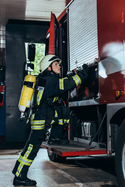 side view of female firefighter with fire extinguisher on back closing truck at fire department