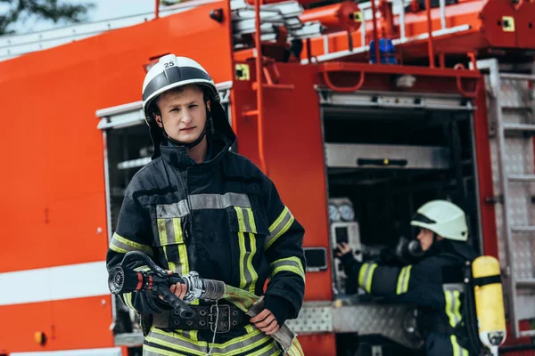 Bombeiro Masculino Uniforme Segurando Mangueira Água Enquanto Colega Verificando Equipamentos — Fotografia de Stock