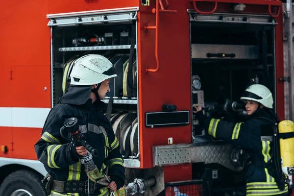 Пожарный Форме Держит Водяной Шланг Пока Коллега Проверяет Оборудование Грузовике — стоковое фото