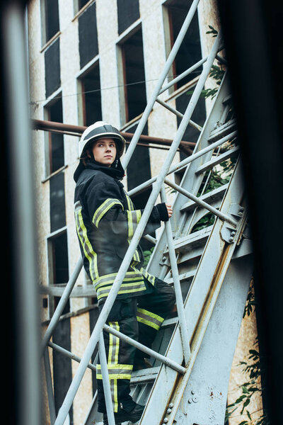 female firefighter in protective uniform and helmet standing on ladder on street