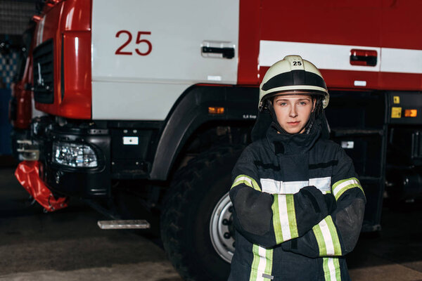 portrait of female firefighter in helmet with arms crossed standing at fire station with truck behind