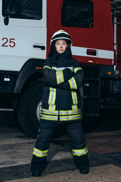 female firefighter in helmet with arms crossed standing at fire station with truck behind