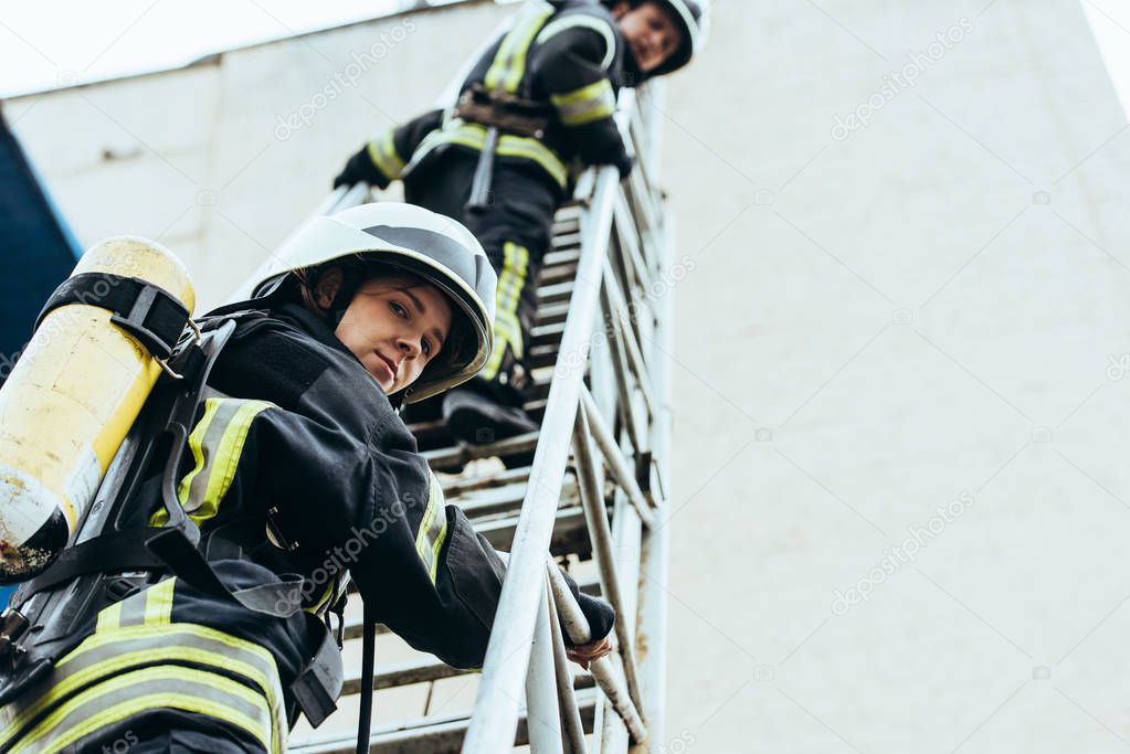 low angle view of firefighters in helmets standing on ladder and looking at camera