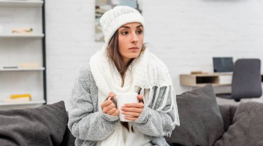 freezed young woman in warm clothes holding cup of warming tea while sitting on couch at home clipart