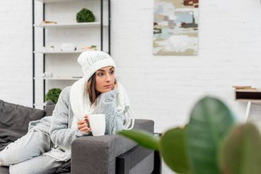 thoughtful young woman in warm clothes holding cup of hot tea and looking away while sitting on couch clipart