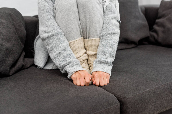 cropped shot of freezed woman in warm clothes sitting on couch and warming her feet