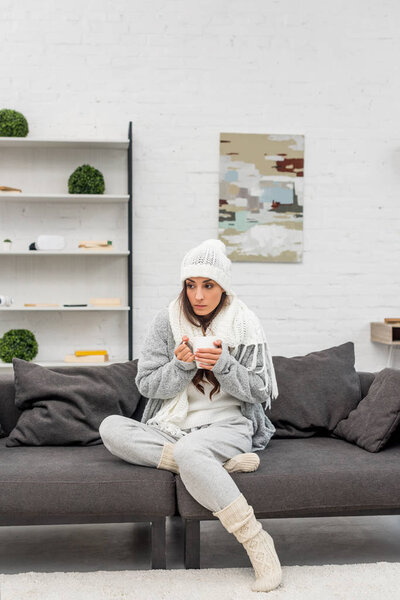 freezed young woman in warm clothes holding cup of hot tea while sitting on couch at home