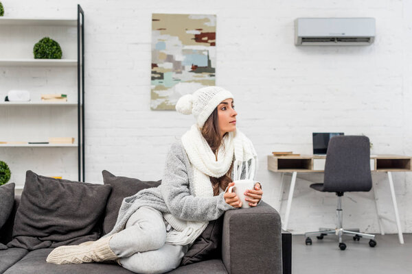 thoughtful young woman in warm clothes holding cup of hot tea and looking away while sitting on couch at home