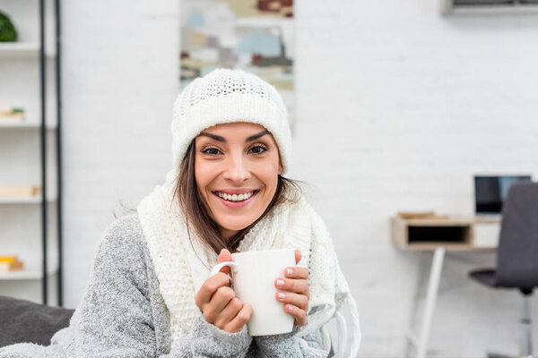 улыбающаяся молодая женщина в теплой одежде держит чашку горячего чая дома
