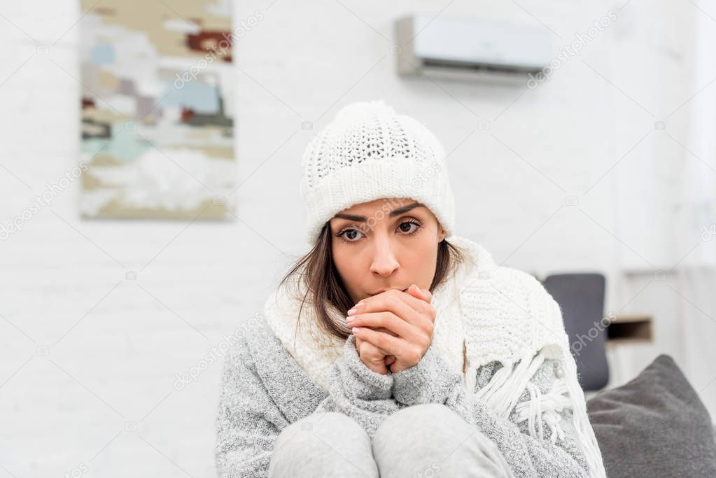 freezed young woman in warm clothes sitting on couch at home and blowing at hands