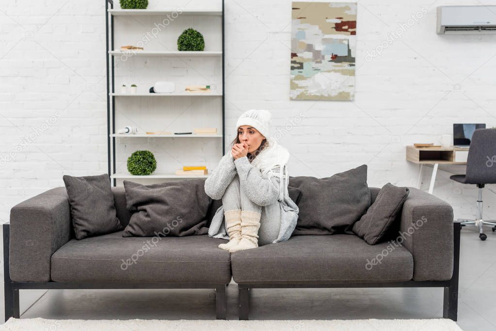 freezed young woman in warm clothes sitting on sofa at home