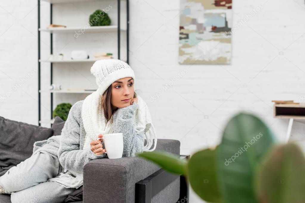 thoughtful young woman in warm clothes holding cup of hot tea and looking away while sitting on couch