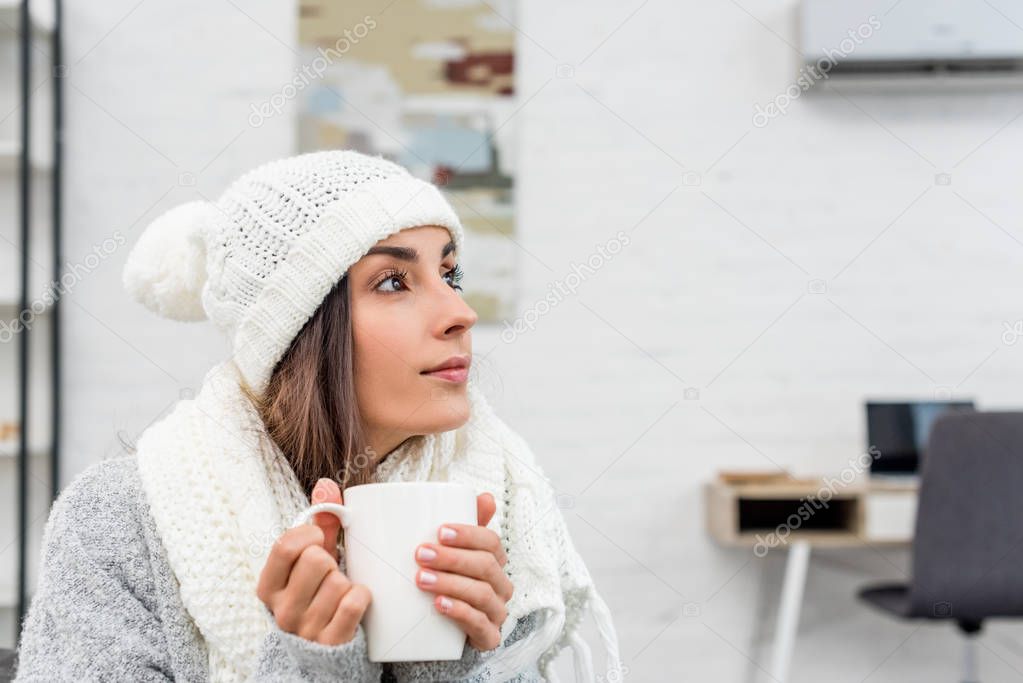 thoughtful young woman in warm clothes holding cup of hot tea at home and looking away