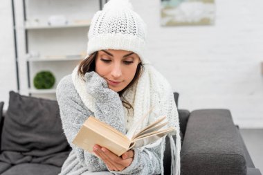 young woman in warm clothes sitting on couch at home and reading book