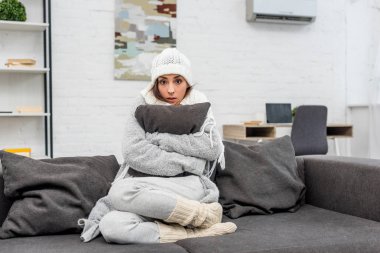 freezed young woman in warm clothes sitting on couch and hugging cushion at home clipart