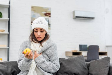 sick young woman in warm clothes holding lemon at home clipart