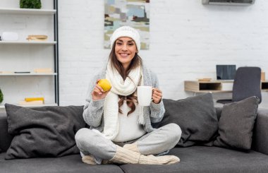 happy young woman in warm clothes holding lemon and cup of tea at home clipart