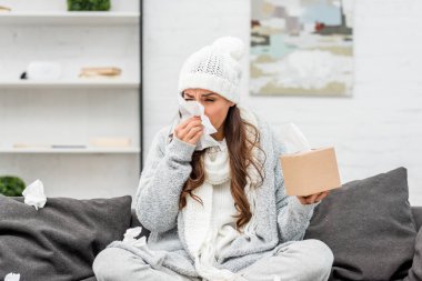 diseased young woman in warm clothes sitting on messy couch and sneezing with paper napkins at home clipart