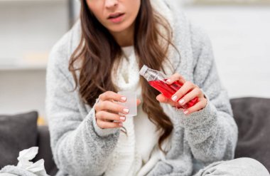 cropped shot of sick young woman in warm clothes pouring red cough syrup while sitting on couch at home clipart