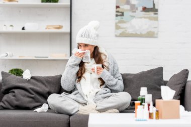 sick young woman in warm clothes sneezing while sitting on couch at home clipart
