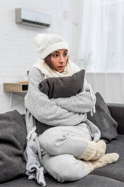 freezed shocked woman in warm clothes sitting on couch and hugging cushion at home