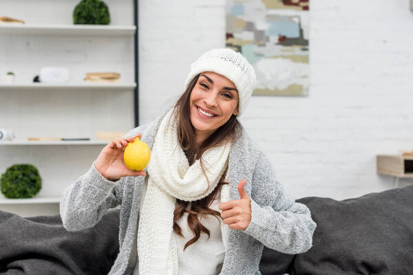 smiling young woman in warm clothes holding lemon and showing thumb up