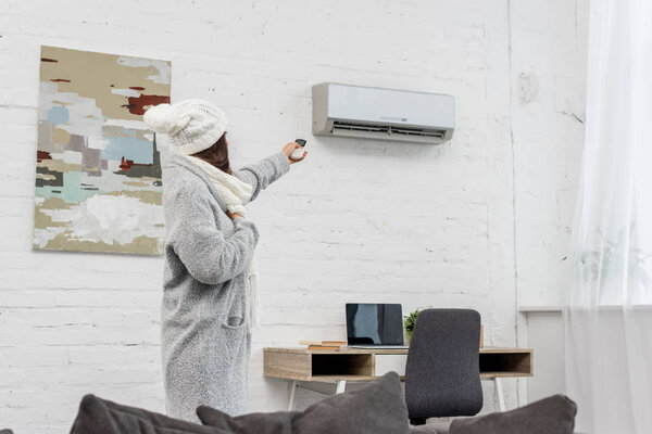 beautiful young woman in warm clothes pointing at air conditioner with remote control at home