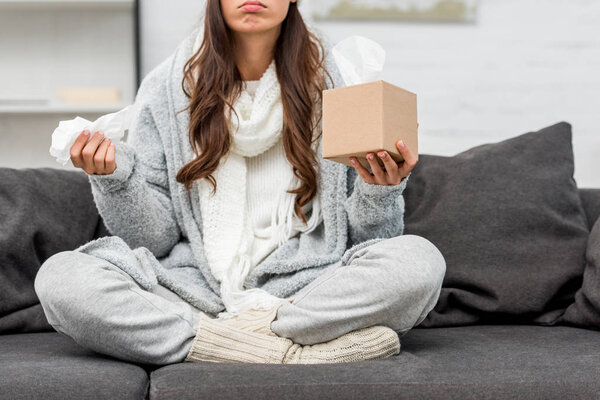 обрезанный снимок больной молодой женщины в теплой одежде, сидящей дома на диване и держащей коробку бумажных салфеток
