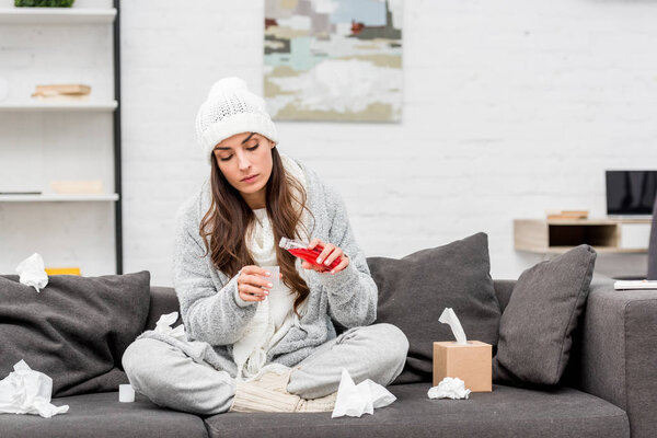 diseased young woman in warm clothes pouring red cough syrup while sitting on couch at home