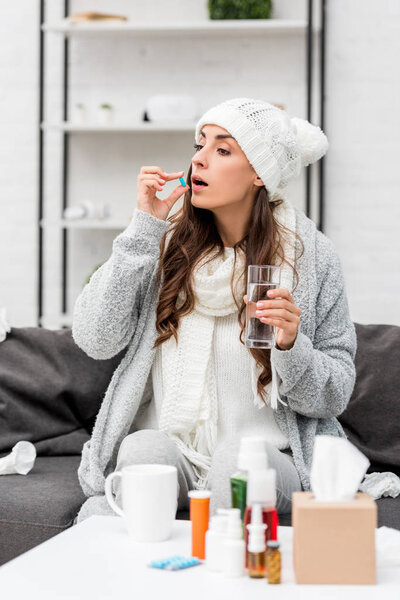 больная молодая женщина в теплой одежде принимает таблетки на диване дома
