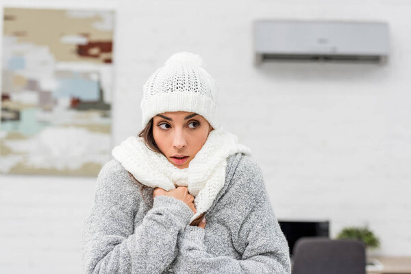 close-up portrait of freezing young woman in warm clothes with air conditioner on background