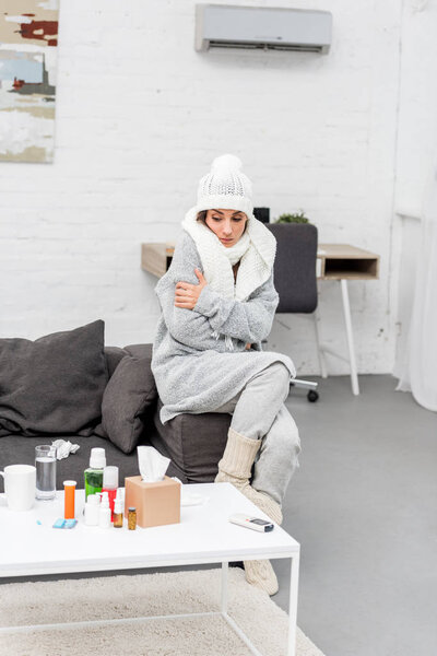 sick young woman in warm clothes sitting on couch and looking at medicines on table