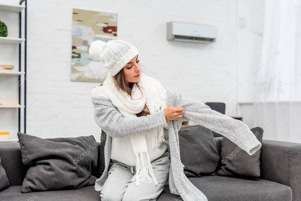 freezed young woman putting on warm clothes while sitting on couch