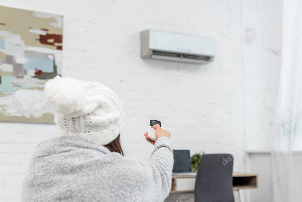 rear view of freezed young woman in sweater pointing at air conditioner with remote control