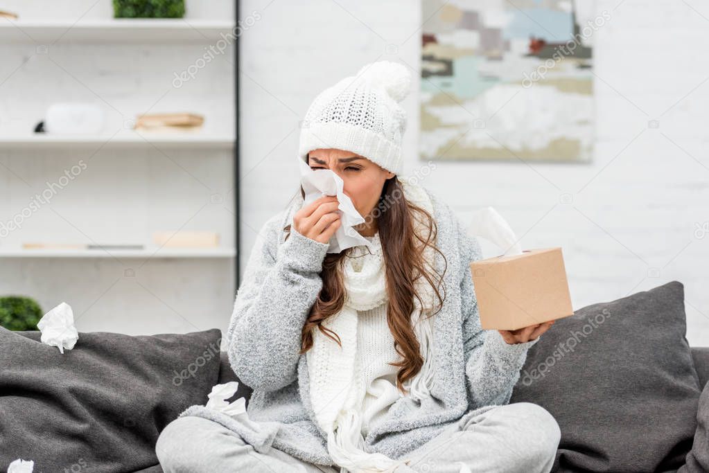 diseased young woman in warm clothes sitting on messy couch and sneezing with paper napkins at home